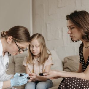 A pediatrician checks a child's blood sugar levels while her mother assists at home.