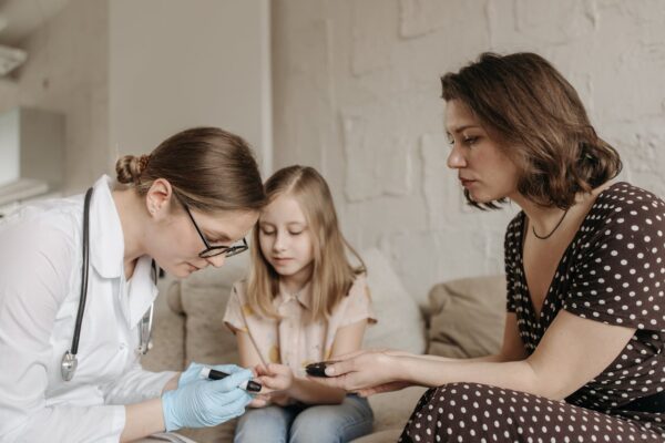 A pediatrician checks a child's blood sugar levels while her mother assists at home.